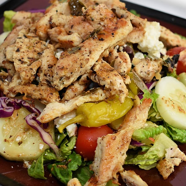 Close-up of a chicken salad with lettuce, tomatoes, cucumbers, red onions, feta, and peppers.