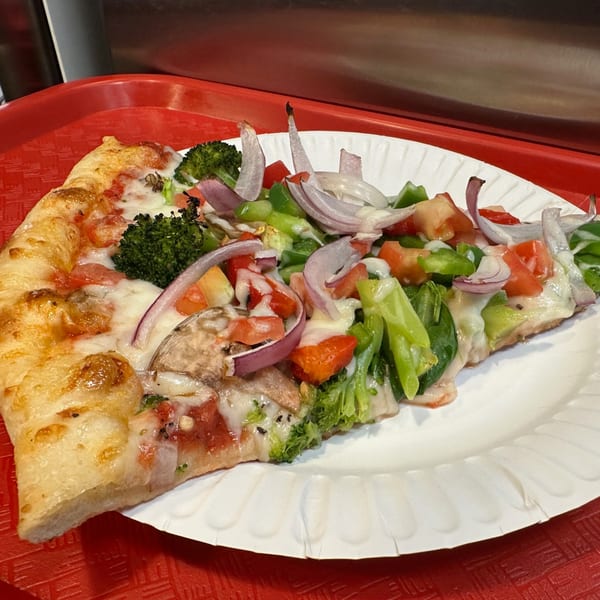 A slice of Eddies Pizza with broccoli, red onions, tomatoes, and green peppers on a white plate.